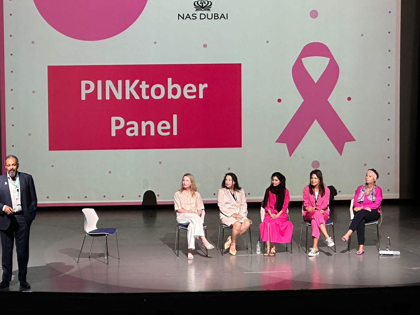 Five panelists seated and a speaker standing on a stage in front of a large screen reading PINKtober Panel, NAS Dubai, with a pink ribbon.